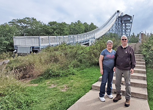 Tom and Patti Kell stand next to the imposing Pine Mountain Ski Jump in Michigan, which at 176 feet is one of the highest artificial ski jumps in the world. “Scary to even be on the ground imagining the jump,” Patti observed.  The jump is located across the river from the small town of Aurora, Wisc.