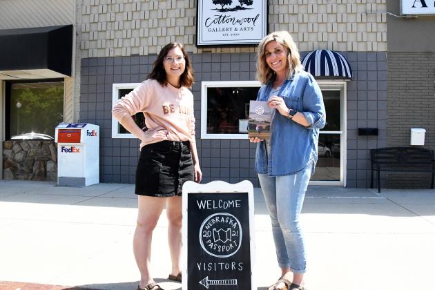 Tori Swanson (left) and Desiree Christenson show off both the Nebraska Passport booklet and their sign welcoming visitors to the Cottonwood Gallery and Arts.