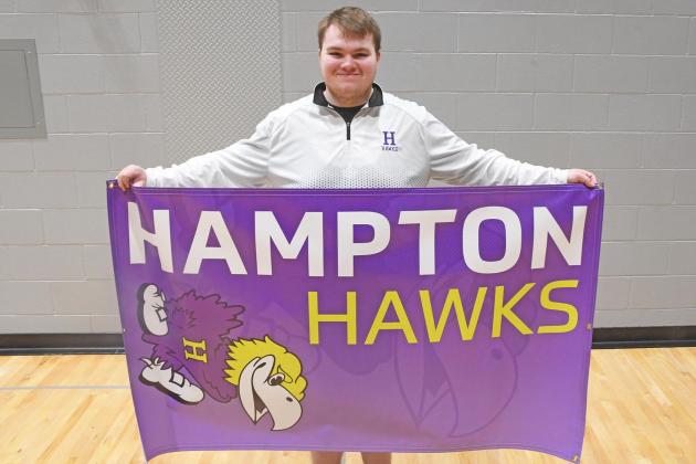 Michael McFarlane stands with his banner for the Hampton Hawks. Each banner has the school name, a small mascot in the foreground and a transparent mascot in the background.