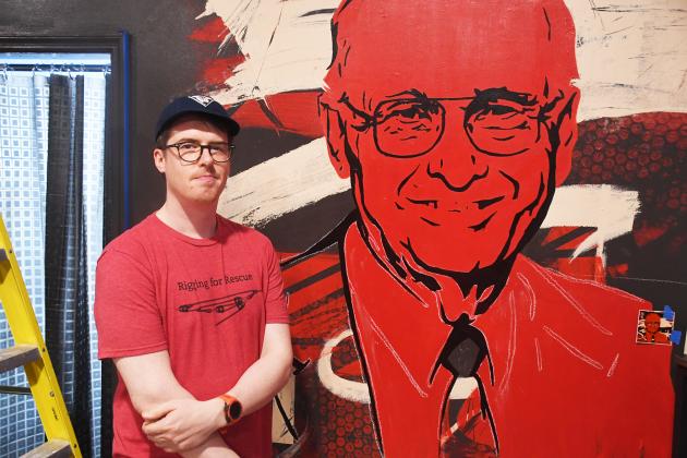 Artist Brandon Marr stands next to the mural of Merwyn Davidson.  Marr had used the design for a sticker made around the time of Davidson’s passing painted mostly in vibrant red and black outline.