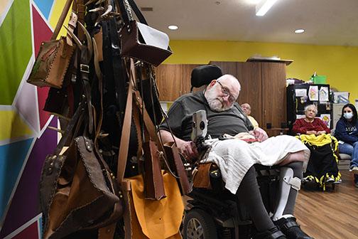 Leather crafter Curtis Marlatt poses next to a rack of bags that he has made at Westfield Quality Care in Aurora as other residents and workers look on. Marlatt makes leather items for residents, family and others in a hobby that keeps him engaged at the care facility.