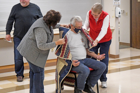 Art Van Luchene (left) is wrapped in his Quilt of Valor during the ceremony held up by Jim Willis and Claire Frevert. Jeanette Friesen gives an explanation of the meaning of the quilt.