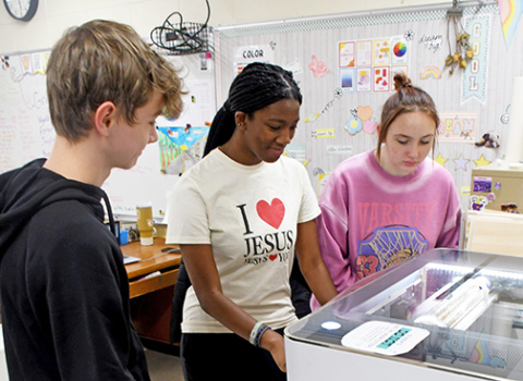 Hampton senior art students, from left, Lofton Ferguson, Taryn Stettner and CoralAnn Van Landingham watch the art department’s new Glowforge laser engraver as it cuts designs into slate coasters. 