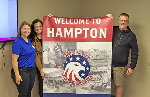 Jana Gibson, left, and Haley Bamesberger of the Hamilton County Nebraska Celebrate America 250 Committee present a commemorative banner to the Village of Hampton Monday night. Accepting the banner on behalf of the village is village board chairman James Parsley. 