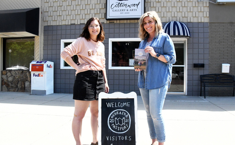 Tori Swanson (left) and Desiree Christenson show off both the Nebraska Passport booklet and their sign welcoming visitors to the Cottonwood Gallery and Arts.