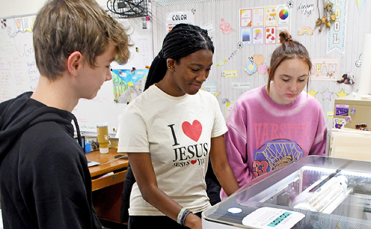 Hampton senior art students, from left, Lofton Ferguson, Taryn Stettner and CoralAnn Van Landingham watch the art department’s new Glowforge laser engraver as it cuts designs into slate coasters.
