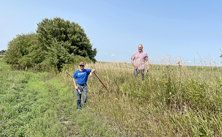 Two friends of David Meyer pose on Nebraska farm land to illustrate the impact of erosion and topsoil loss over many years. “These two fields were once the same level across the field fence; it is a striking difference,” Meyer said. The man on the right stands on unplowed prairie while the man on the right stands on land that has been in pasture for the last 20 years, but previous to that had been row cropped.