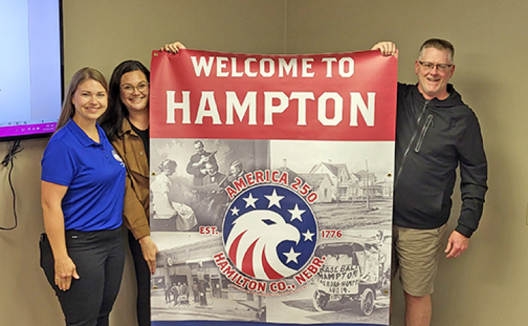 Jana Gibson, left, and Haley Bamesberger of the Hamilton County Nebraska Celebrate America 250 Committee present a commemorative banner to the Village of Hampton Monday night. Accepting the banner on behalf of the village is village board chairman James Parsley.