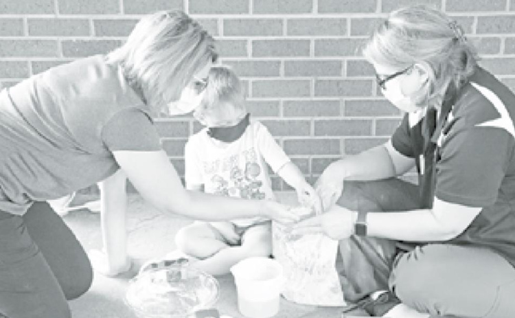 MCHI Ocupational Therapist Kim Eberly (left) helps young Knox Sabata explore his sandy activity. MCHI Occupational Therapist Carla Christenson also encouraged the young Auroran to squish the material between his fingers. News-Register/Cheyenne Rowe